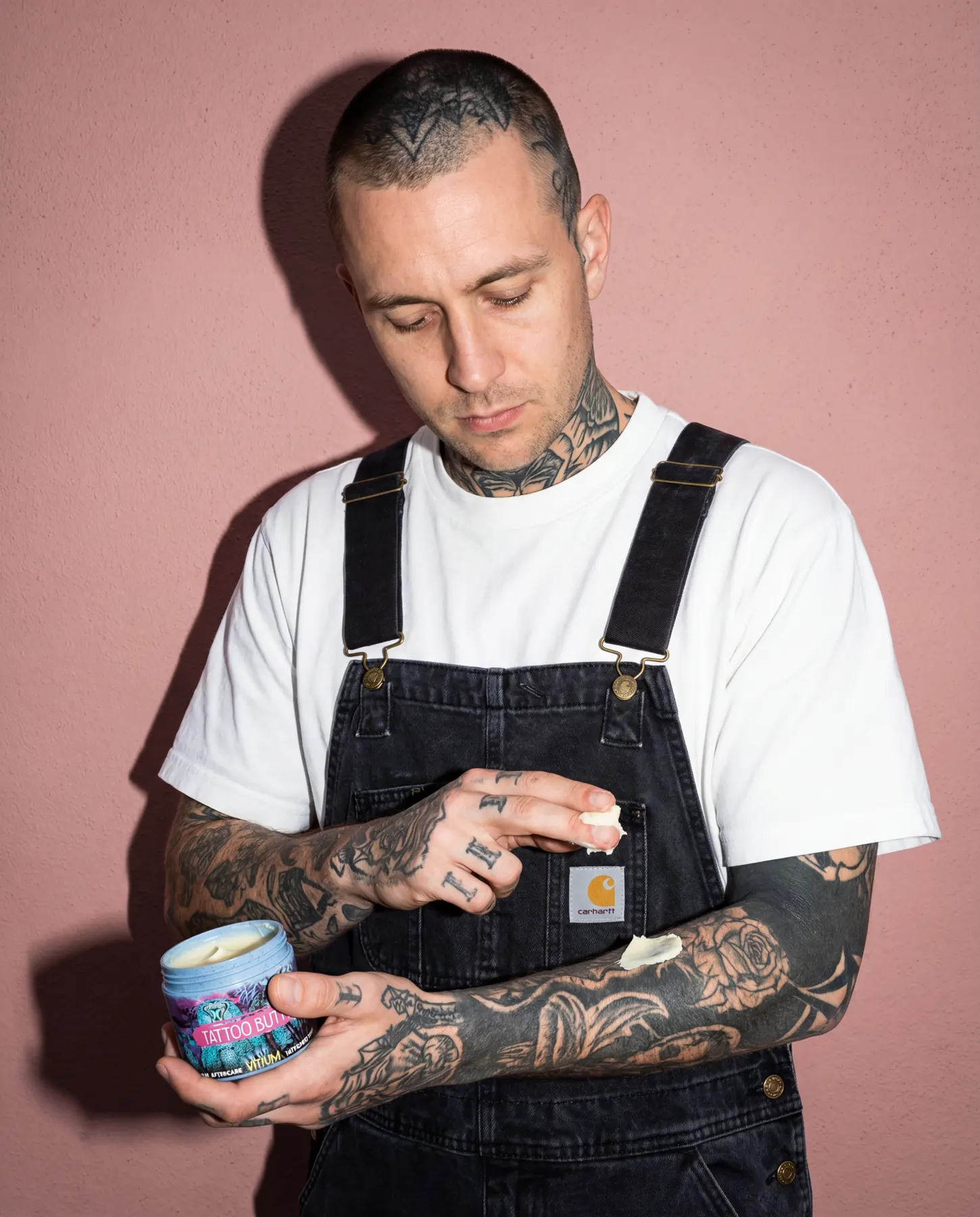 Heavily tattooed artist in Carhartt overalls applying VITIUM Tattoo Butter to a blackwork tattoo on his forearm, against a dusty pink wall