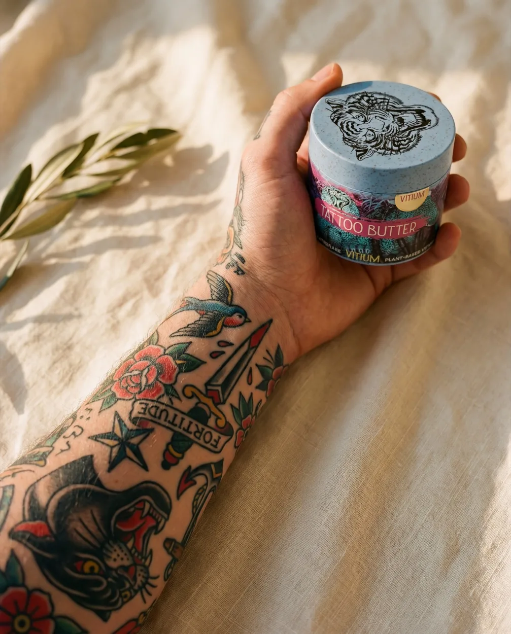 Tattooed arm holding VITIUM jar on sunlit linen with olive branch